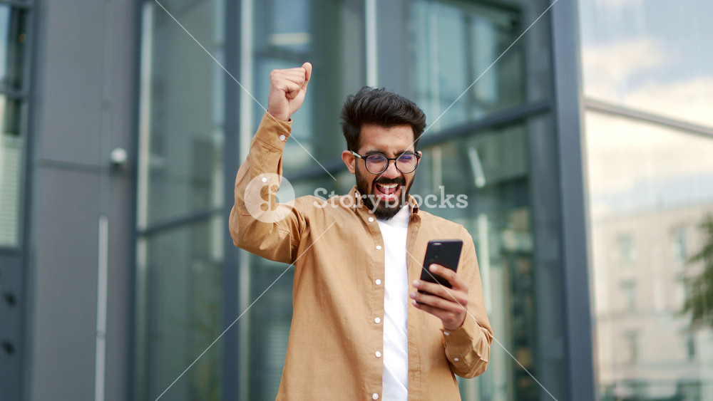 Excited man celebrates victory after receiving great news on smartphone while standing on street near office building. Happy handsome businessman holding hand in triumph gesture showing yes Wow effect