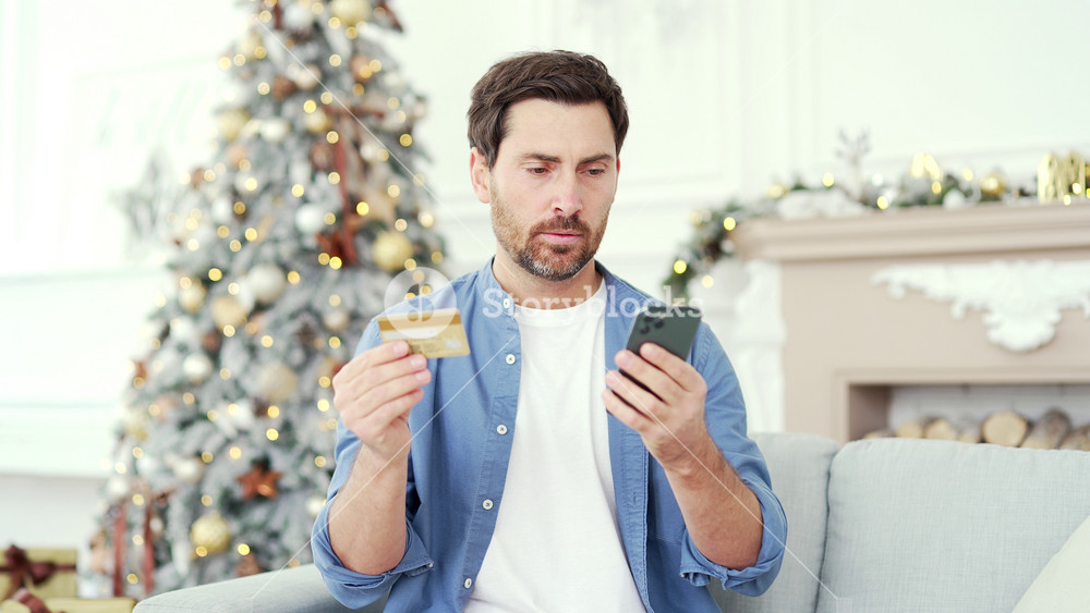 Frustrated man discovered fraud entering credit card number on smartphone sitting at home during winter New Year Christmas Xmas holidays. Upset male saw that money had been stolen from his account