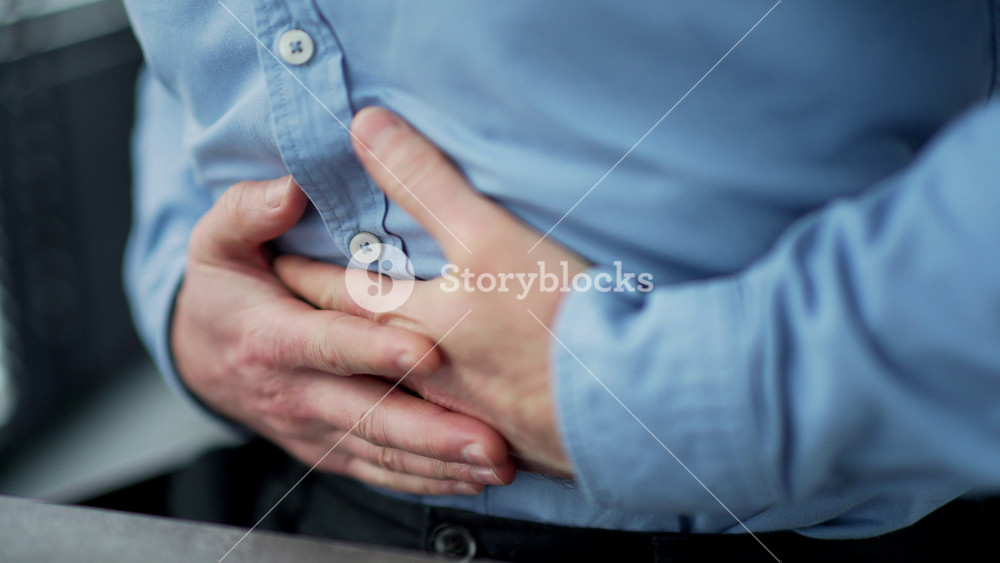 Close up. Male hands holding massaging stomach. Businessman in a blue shirt feels stomach pain sitting at workplace in office. Sick man suffers from bloating, gastritis poisoning or spasms