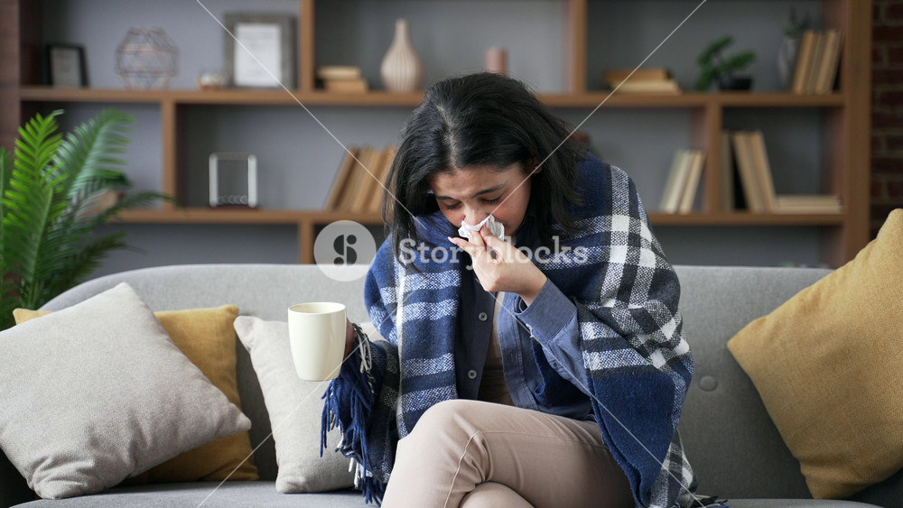 Sick young woman suffering from cold and flu sitting on sofa in living room at home. An unhealthy adult female wrapped in a blanket coughs, wipes her nose with a handkerchief and drinks hot tea