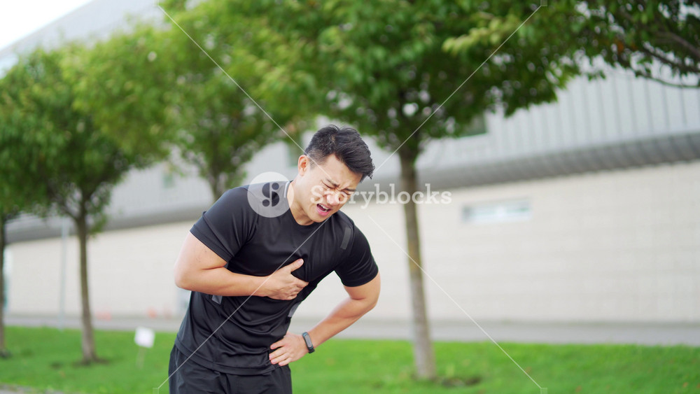 young adult asian athlete while jogging chest injury, severe heart attack pain and shortness of breath, male runner, signed heartbeat and high blood pressure while exercising in a city park. outdoors