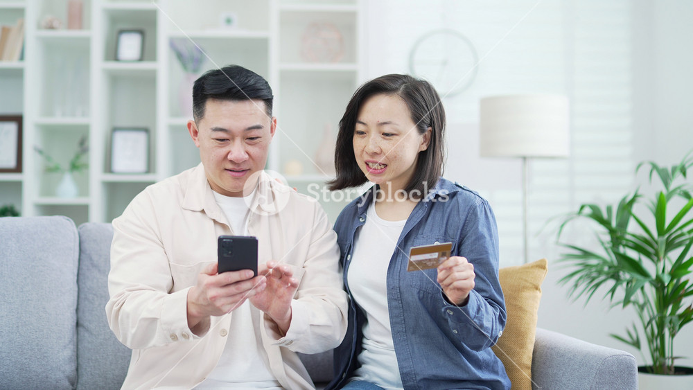 Happy asian couple doing online shopping by entering credit card number on smartphone while sitting on sofa at home. Smiling husband and wife are satisfied with the purchase and the promotional price