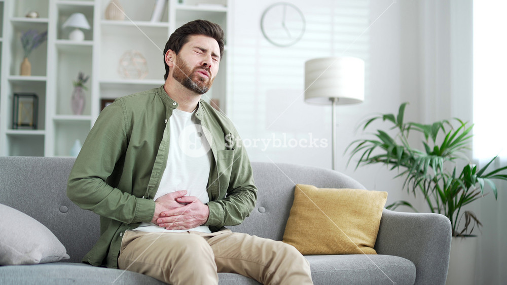 Sick man feels stomach pain while sitting on sofa in living room at home. Upset handsome bearded male has heartburn, gastritis, bloating or poisoning. Male suffers from spasms and constipation