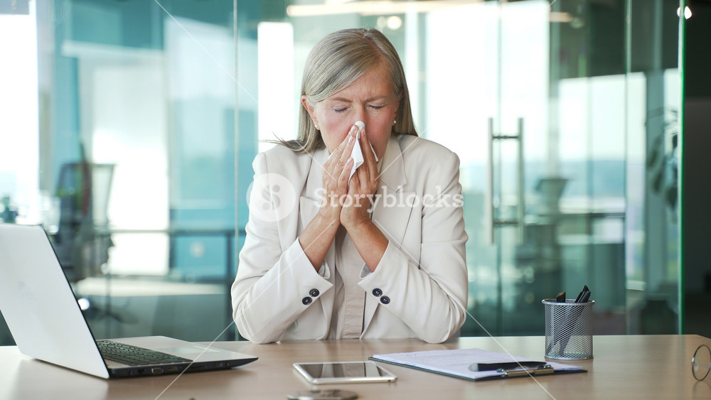 Sick senior gray haired businesswoman suffering from seasonal allergies sitting at a workplace at a desk in a business office. Allergic lady entrepreneur sneezes and wipes her nose with a handkerchief
