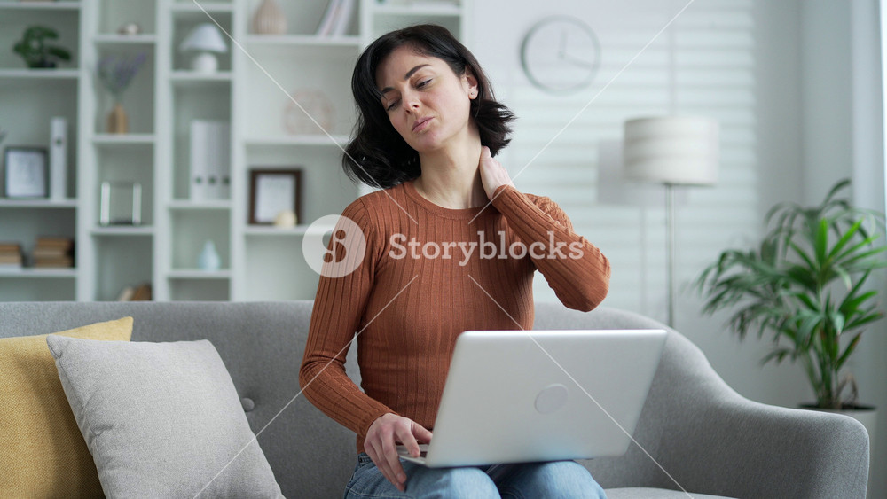 Tired young adult woman suffering from neck pain while working on a laptop while sitting on the sofa in the living room at home. Upset woman massages and rubs sore muscles with her hands, stretches