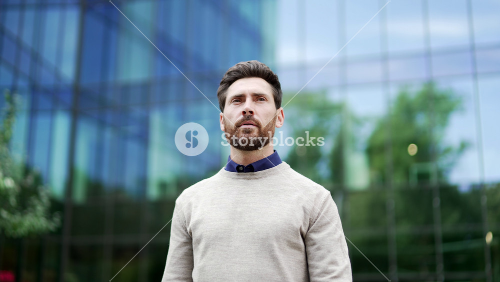 Confident businessman standing near modern glass office building. Professional appearance and thoughtful expression convey success. Successful man entrepreneur walks to business district of the city