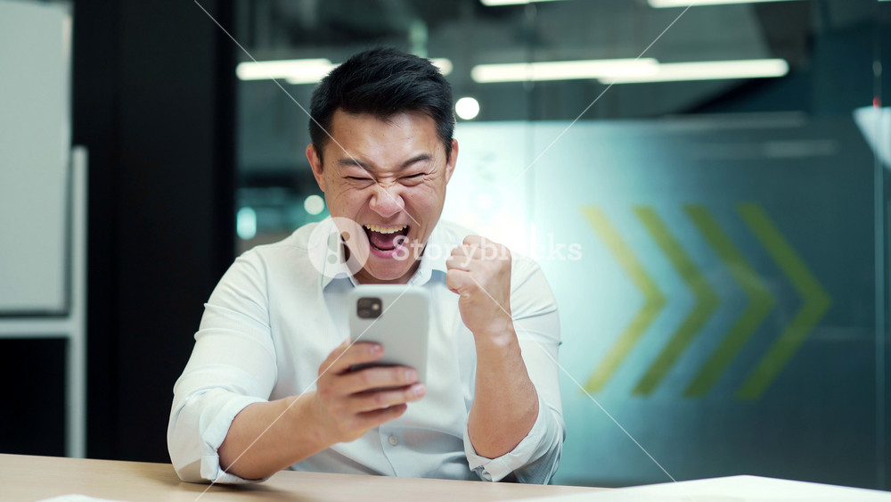 Happy asian man employee enjoys success read good news in smartphone. businessman win mobile online bet bid money financial prize victory receive offer opportunity on smart phone in the office indoor