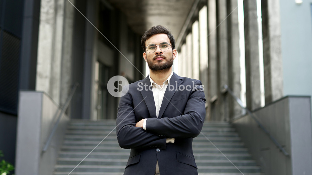 Portrait of serious businessman in formal suit standing on city street near office building. Proud bearded handsome entrepreneur with crossed arms looking at camera. Confident manager posing outdoors