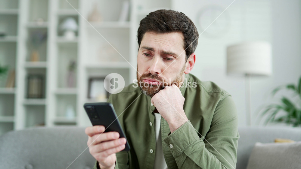 Lonely sad handsome man bored using smartphone sitting on sofa in living room at home. Tired bearded male is depressed and in a bad mood, browses social media on a mobile phone. Close up