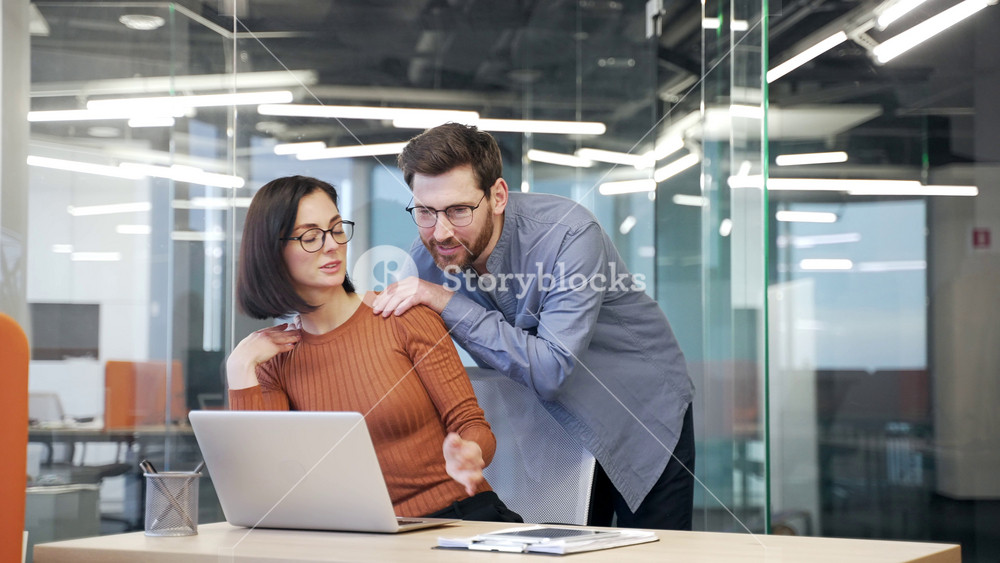 Businessman harasses a young female coworker as places his hands on her shoulder at workplace in business office. Male employee flirts with a shocked worried female colleague.