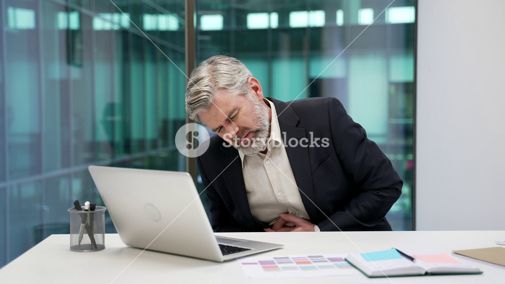 Unhealthy mature businessman feels stomach pain sitting at workplace in business office. Sick worker in formal suit has heartburn, gastritis or poisoning. Male suffers from spasms or constipation