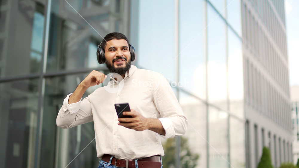 Happy joyful handsome employee listens to music on wireless headphones while walking on the street near the office building. Smiling man turns on the playlist on his mobile phone and enjoys the song