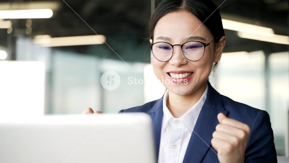 Happy young asian businesswoman reading great news on laptop sitting at workplace in business office. Excited woman worker celebrates success, rejoices at the win, smiles, shows gesture yes. Close up