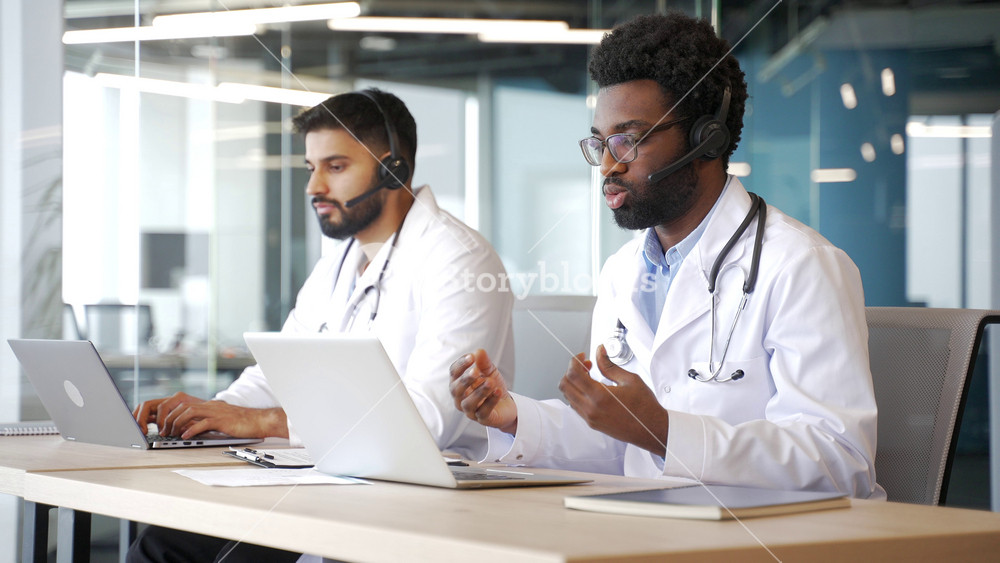 Two male doctors in white coats with headsets working on laptops in medical call center. Multiethnic healthcare professionals helping patients remotely, offering consultation and advice in office.