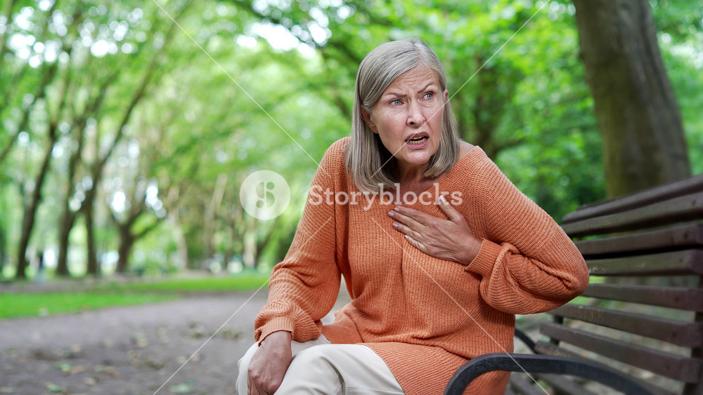 Sick senior female with gray hair suffering from chest tension, heart attack sitting on bench in city park. Elderly woman feels anxious, holding her chest with her hand, needs urgent medical attention