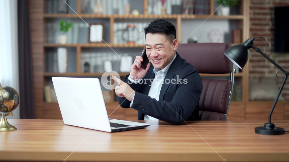 Satisfied Asian business man talking on a mobile phone while sitting in the office. Great result, happy manager businessman. Lawyer or banker employee entrepreneur communicates using laptop. indoor
