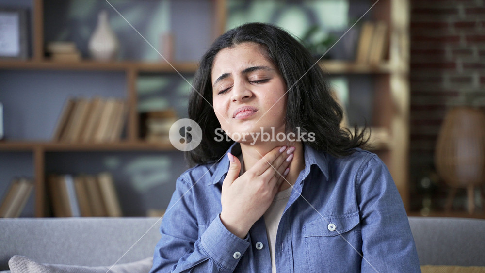 Sick young woman suffering from a sore throat sitting on sofa at home. Unhealthy adult female has cold, virus or flu. Upset girl touching throat with pained expression, massaging sore spot . Close up
