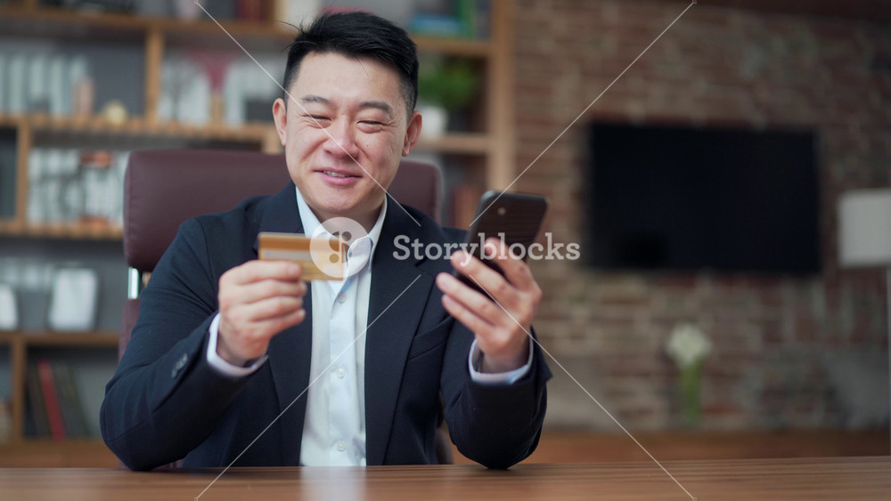 Asian business man shopping online pays by credit card using mobile phone smartphone sitting at desk office, work workplace. Purchase travel, bank service, booking. Male businessman worker in suit