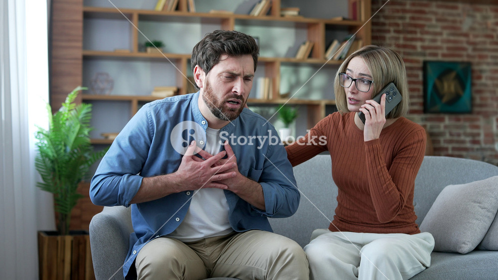 Unwell man having a heart attack while sitting on sofa in room at home. Compassionate wife supports her husband and calls an ambulance by phone. Unhealthy male is breathing hard, holding his chest