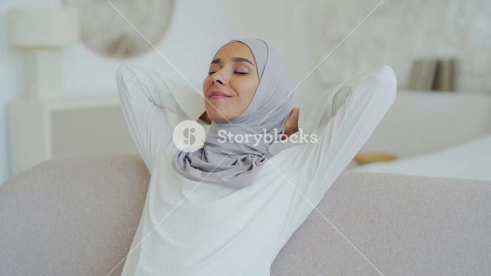 Close up of beautiful young Muslim woman in hijab relaxing on comfortable sofa with hands behind head in the light living room Smiling peaceful girl enjoy calm time at home alone No stress concept