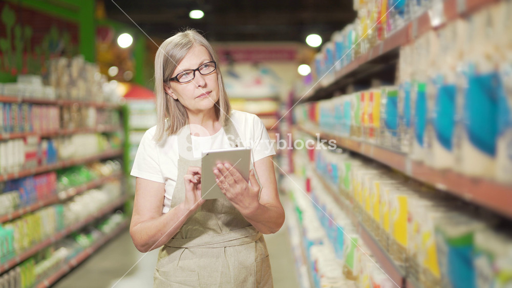 Senior female manager worker in supermarket, grocery store or deli in apron with digital tablet. Mature female small business owner employee inspects shelves inventory products doing checking stock