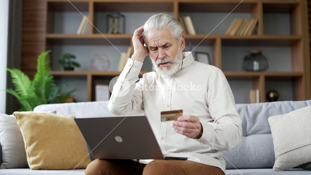 Frustrated elderly senior man discovered fraud while entering credit card number on laptop sitting on sofa at home. Upset mature male became victim of deception, money had been stolen from his account