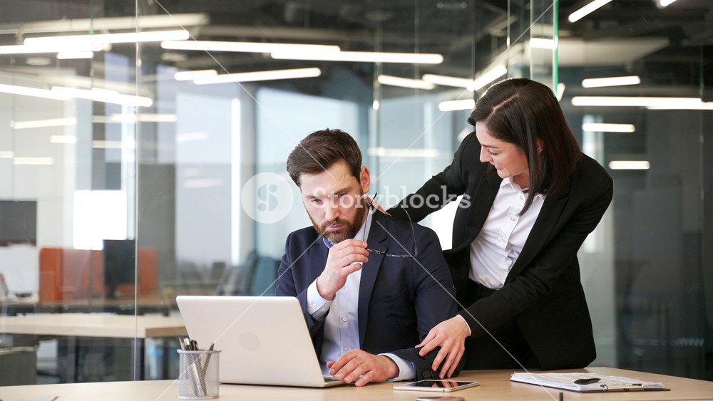 Young businesswoman harasses a male coworker as places her hand on his shoulder at workplace in business office. Female flirts with shocked worried colleague who is working at his desk