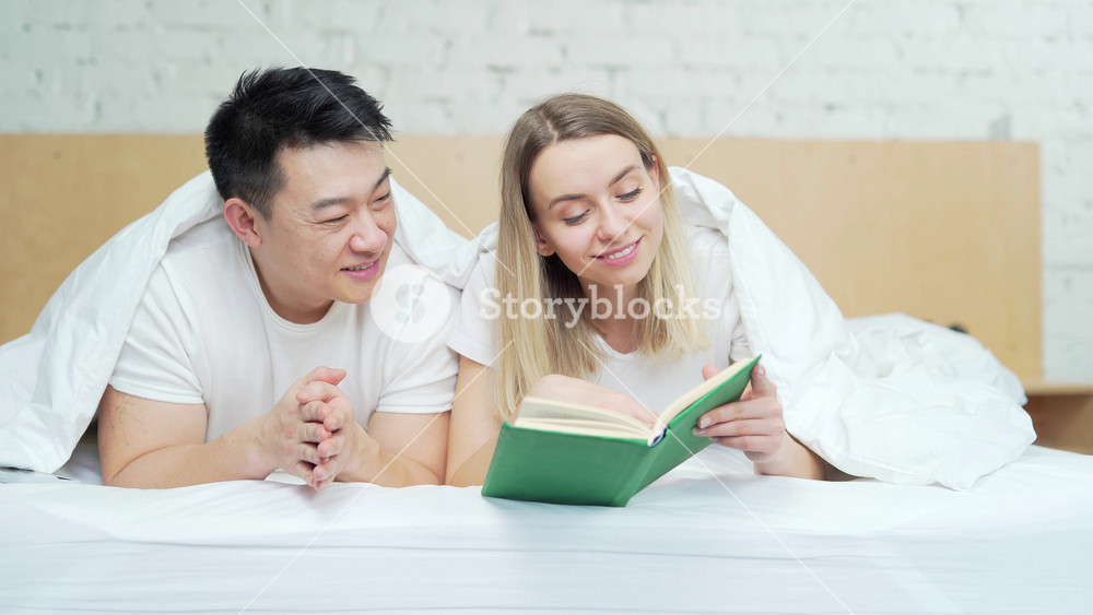 Happy young multiracial family reading a book together lying in bed. Mixed race couple spends leisure time together on weekends in the morning. Cheerful Asian husband and wife in bedroom at home