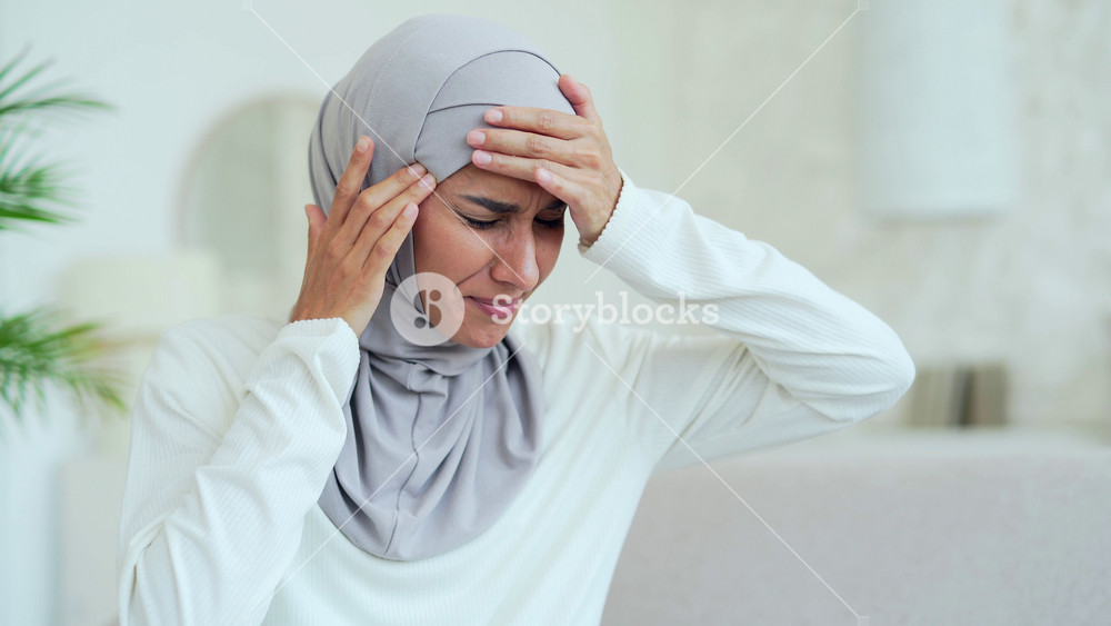 Unhappy young Muslim woman in hijab sitting on couch rubs temple with closing eyes suffering headache painful feelings chronic migraine high or low pressure Stressed upset female feels hurt at home