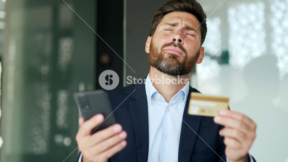 Shocked businessman checks his credit card on smartphone at workplace in business office. Frustrated sad male faced fraud, money was stolen from his account. He became a victim of deception. Close up