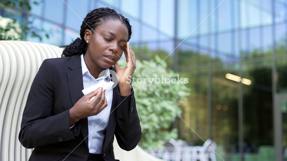 Close-up of an African American businesswoman in a suit suffering from a runny nose near an office building. Coughing and wiping her face with a tissue while sitting on a bench, she feels sick.