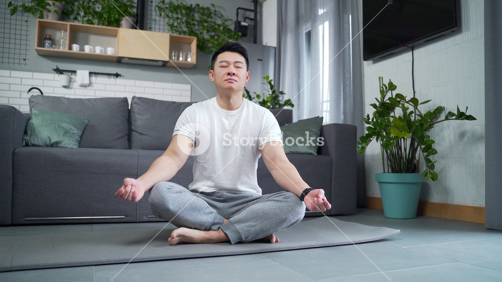 Asian sporty man practicing yoga sitting on mat in lotus position at home in living room. Calm male meditates and relaxes indoors. Fitness sport and healthy active lifestyle. breathes with eyes closed