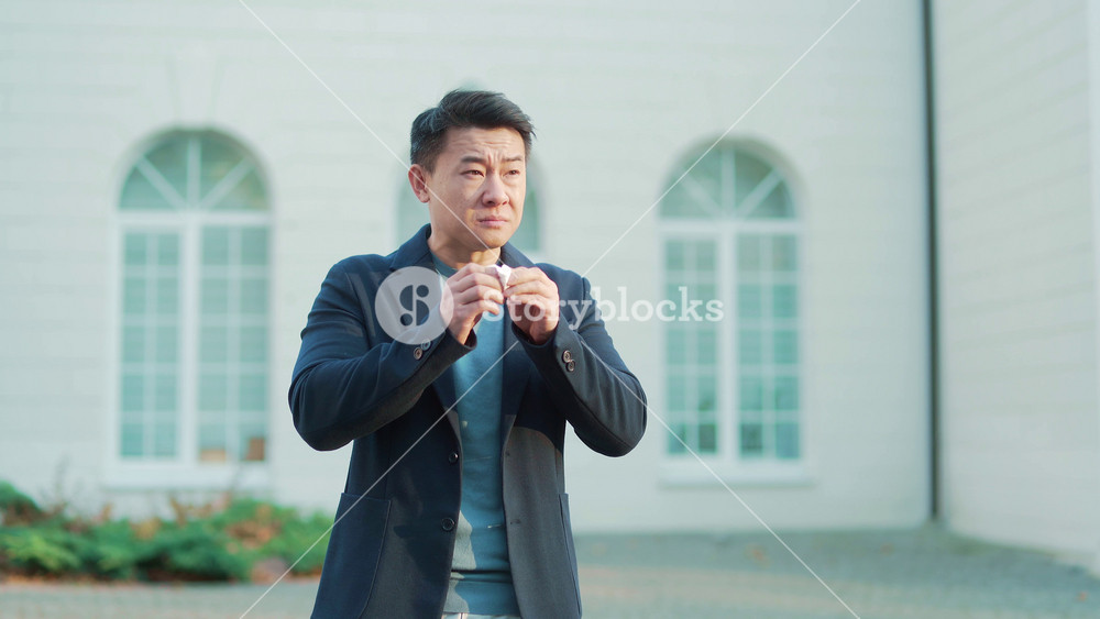 portrait of a sick young asian man who sneezes and coughs uses a handkerchief on the street outdoors. Business Male in a suit suffers from season allergies unhealthy flu while standing outside