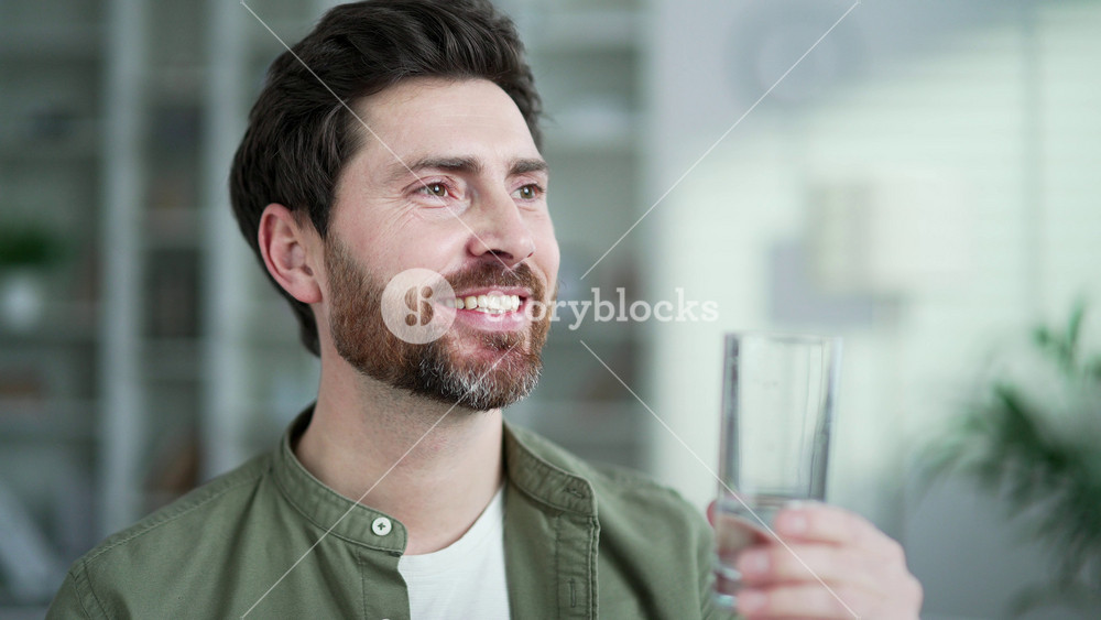 Happy handsome man drinks water from a glass with closed eyes sitting in living room at home. Satisfied smiling bearded male feeling relieved, enjoying clean cool drink, quenching his thirst. Close up