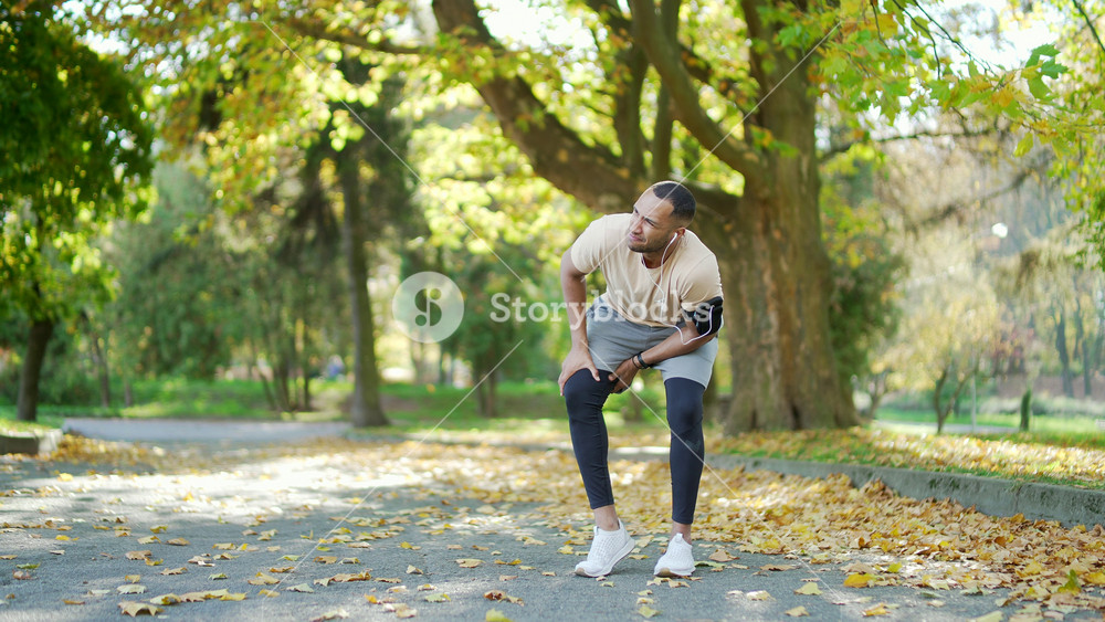 Young male runner experiences muscle pain and discomfort during a jog in the city park. He is massaging and stretching to alleviate the trauma injury he may have sustained while jogging outdoors
