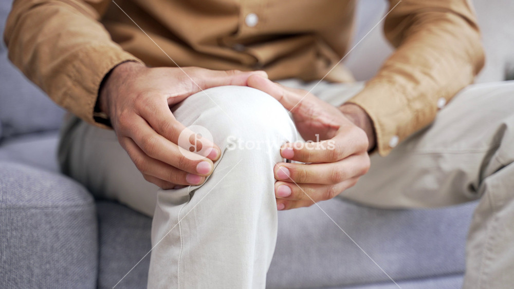 Close up Male hands massaging knee leg muscles while sitting on sofa in living room at home. Sad man in a shirt suffers from painful severe knee joint pain, could not walk, illness, cramps, rheumatism