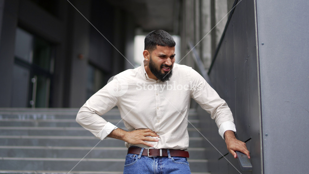 Adult employee experiences a sharp pain in his right side walking down the stairs of an office building. The male has a liver bolt. Sad bearded businessman feels stomach pain, suffers from convulsions