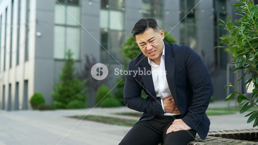 Upset asian businessman in formal suit feels stomach pain sitting on a bench on street near an office building. Sick male has heartburn, gastritis or poisoning, suffers from overeating or constipation