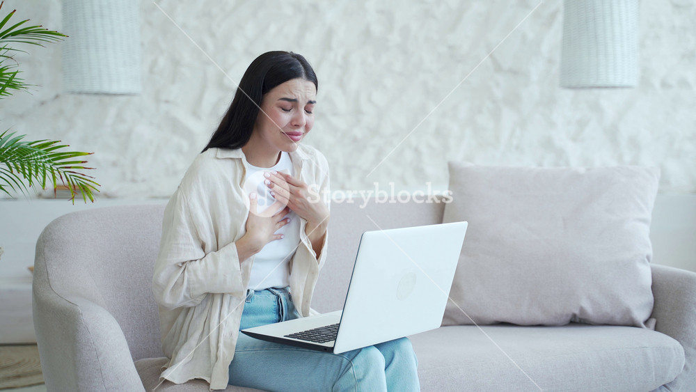 Crying young woman talk with psychotherapist at online counselling session sitting on the couch in the light living room. Depressed female discussing problem with psychological support by laptop.