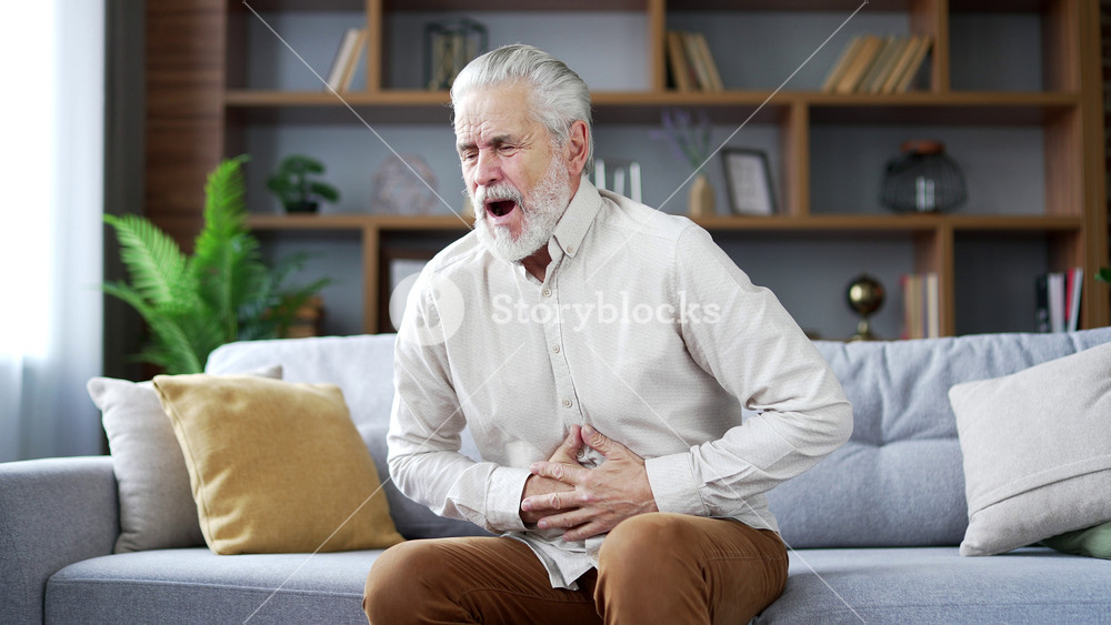 Elderly senior man with gray hair feels stomach pain sitting on sofa in living room at home. Sick adult mature male has heartburn, gastritis or poisoning. Pensioner suffers from spasms or constipation