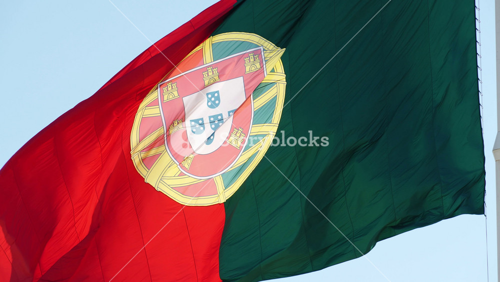 The official coat of arms and armillary sphere of a large Portuguese flag billowing against a light blue sky near Edward VII Park in Lisbon