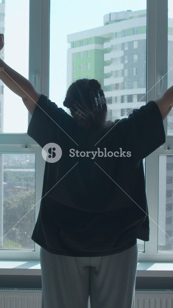 Rear view of a girl in casual clothes in modern bedroom in the morning. Media. Woman looking through the window, raising hands and