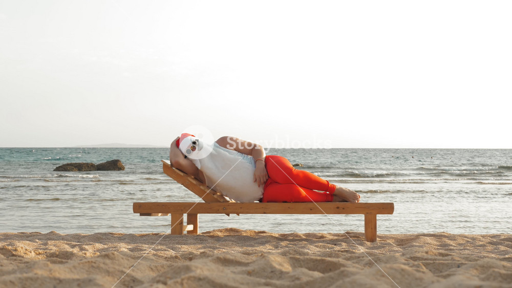 Sunbathing Santa claus relaxing on beach chair near turquoise water. Idea of tropical christmas and quality rest concept.