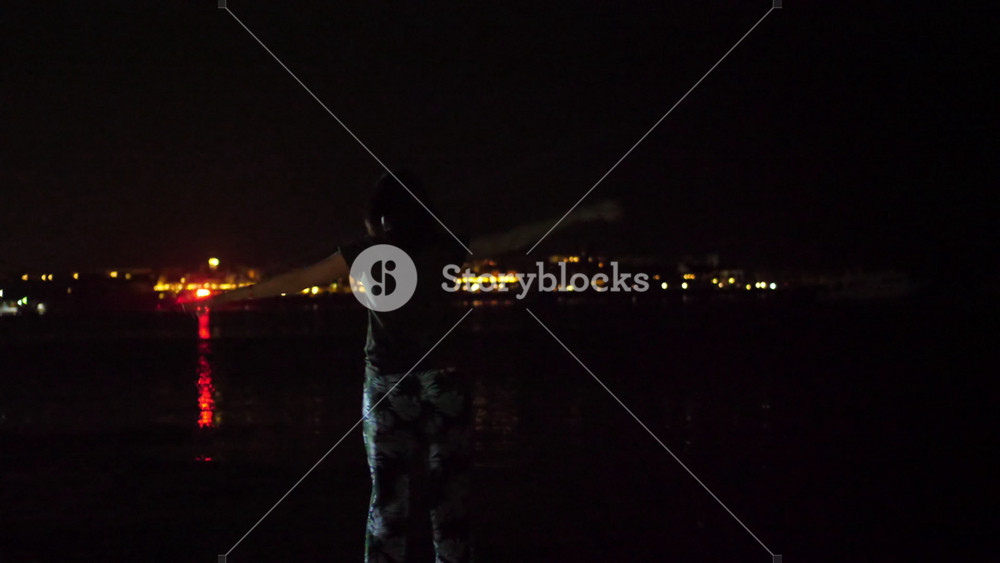 Rear view of young woman dancing against night city lights. Female silhouette enjoying nightlife party atmosphere near water.