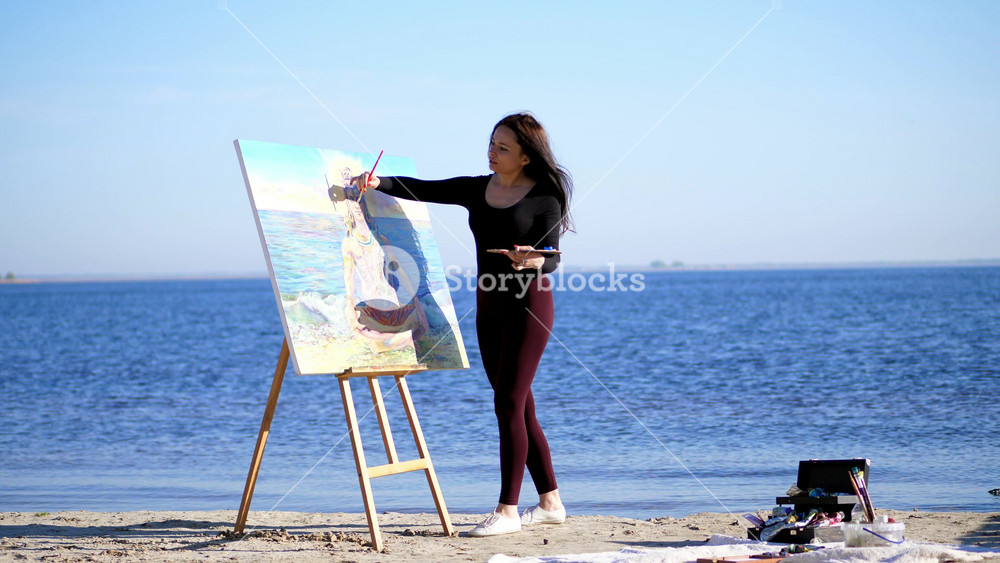 Beautiful woman artist painting God Shiva portrait on easel by river. Spiritual creativity and religion concept at sunrise.