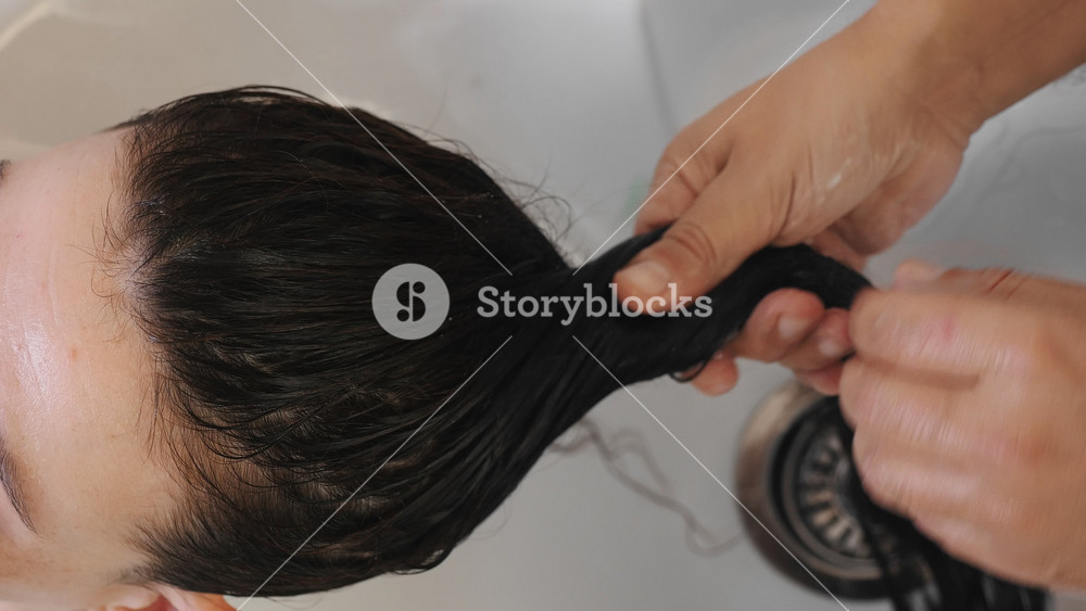 Professional hair care treatment involving rinsing shampoo from brunette woman's head. Stylist using warm water to clean hair in salon wash basin.