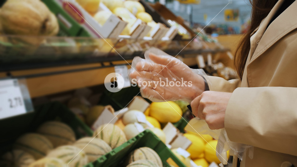 Female shopper putting on blue disposable gloves before selecting fresh produce in a supermarket. Concept of hygiene and health safety in grocery retail.