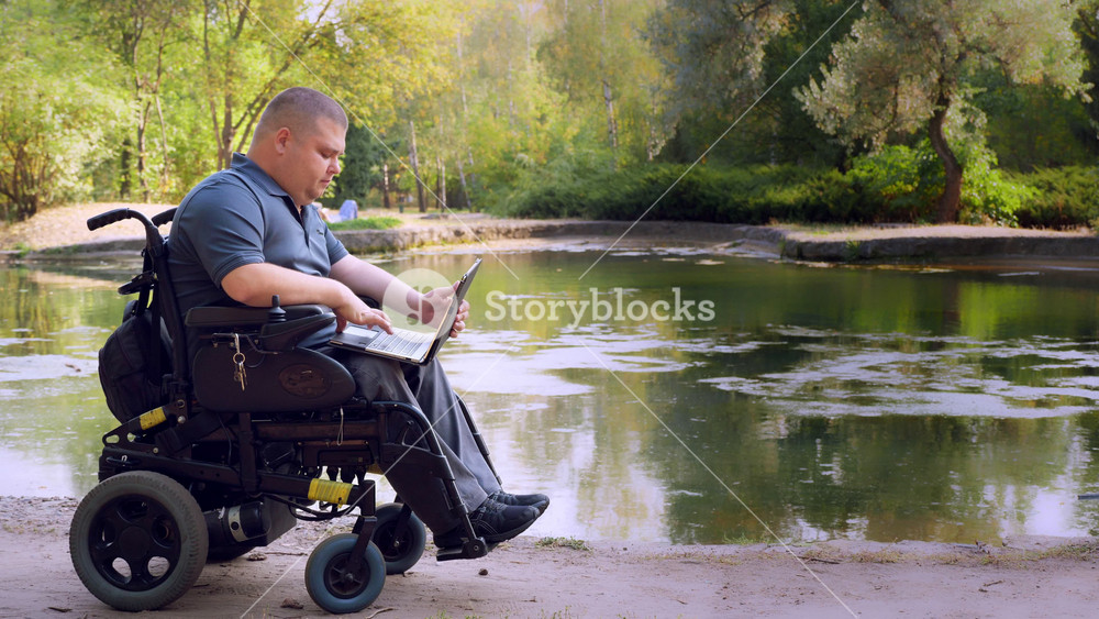 wheelchair man. Handicapped man. young disabled man, sitting in an automated wheelchair and working on a laptop, in a city park, by the lake, on autumn sunny day.