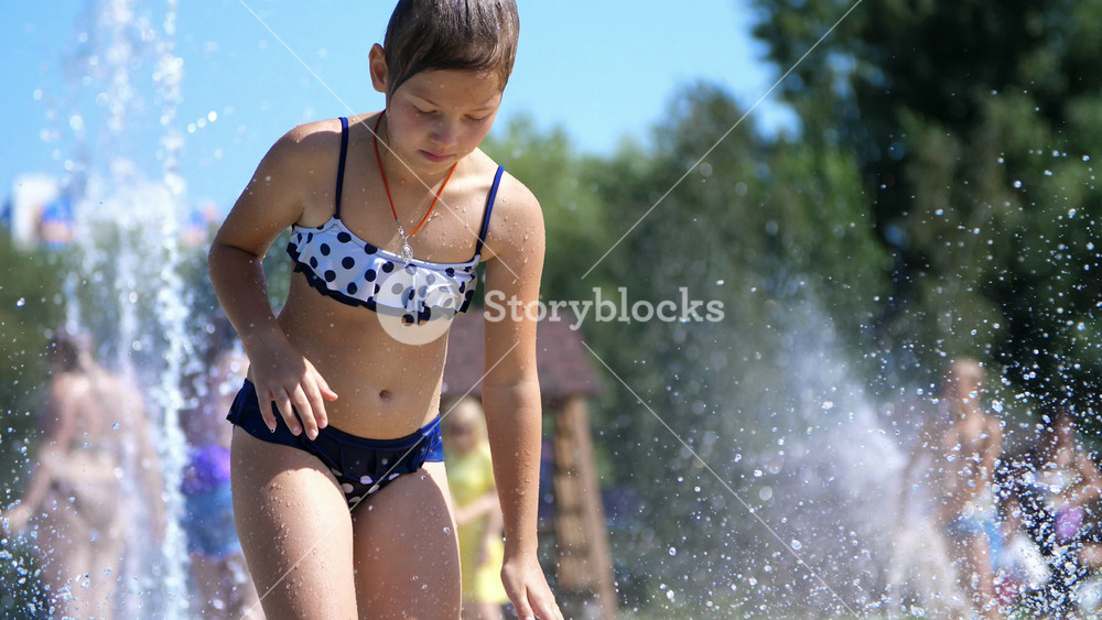 Energetic girl splashing water drops while playing in city fountain. Fun outdoor recreation for kids in hot weather.