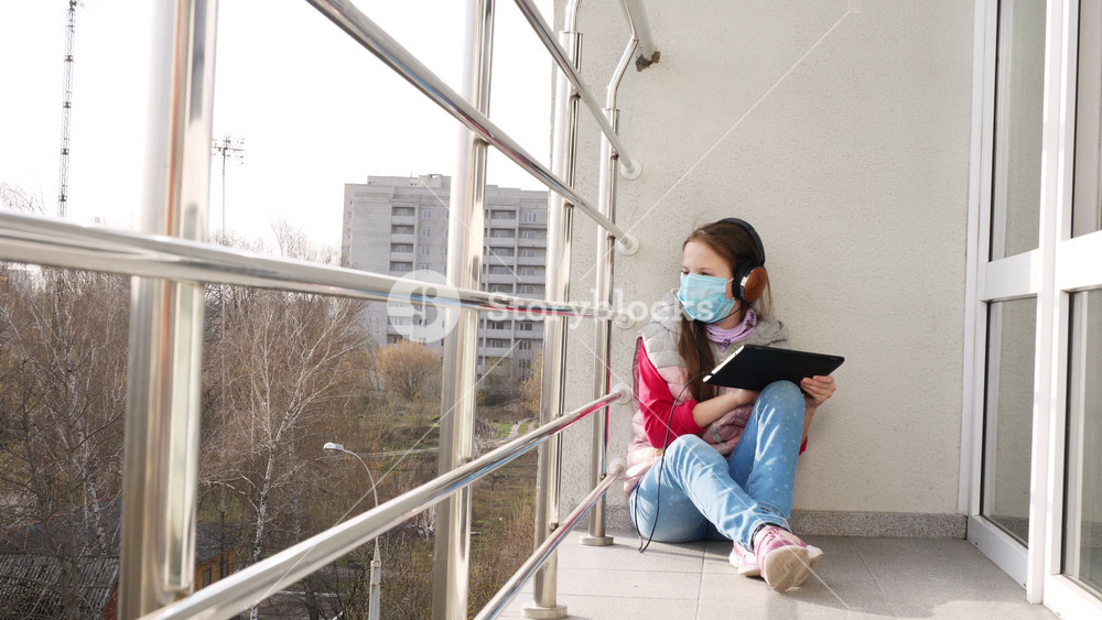 Teenager in medical mask with headphones holding tablet sitting on balcony floor with cityscape in the background. Distance learning during quarantine outdoors.